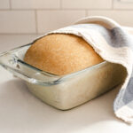 A risen loaf of healthy sourdough bread made with whole wheat flour with a tea towel partially on top.