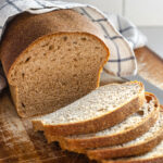 A loaf of healthy sourdough bread on a cutting board with a few slices in the foreground with the loaf in the background with a tea towel on top.