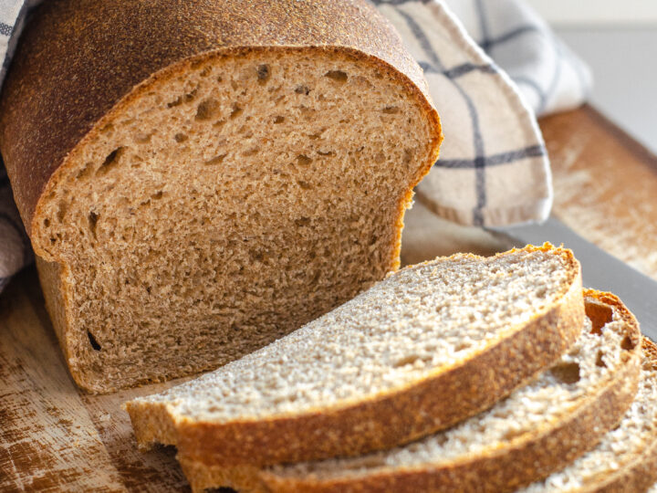 A loaf of healthy sourdough bread on a cutting board with a few slices in the foreground with the loaf in the background with a tea towel on top.