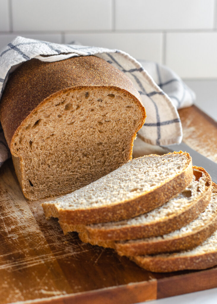 A loaf of healthy sourdough bread on a cutting board with a few slices in the foreground with the loaf in the background with a tea towel on top.