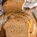 A loaf of healthy whole wheat sourdough sandwich bread on a cutting board with a few slices in the foreground with the loaf in the background with a tea towel on top.