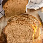 A loaf of healthy whole wheat sourdough sandwich bread on a cutting board with a few slices in the foreground with the loaf in the background with a tea towel on top.