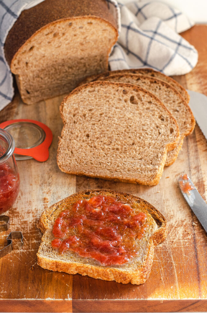 A toasted slice of whole wheat sourdough sandwich bread with butter and jam and more slices of bread in the background, a jam jar to the left, and a knife to the right.
