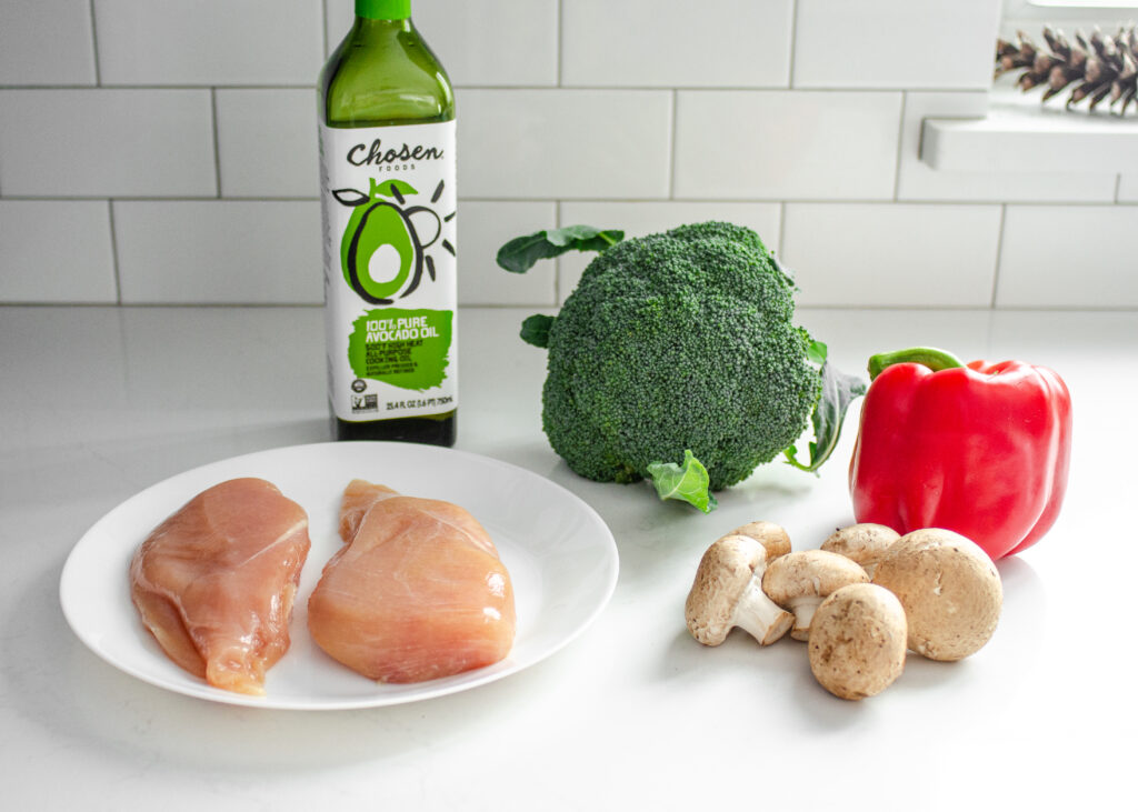 Chicken, avocado oil, broccoli, red bell pepper, and mushrooms on a white countertop with subway tile backsplash. 