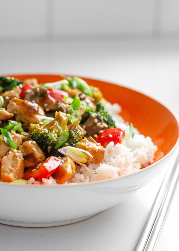 An orange bowl of rice with chicken stir fry on top. Learn how to make my healthy chicken stir fry recipe with these step by step instructions!