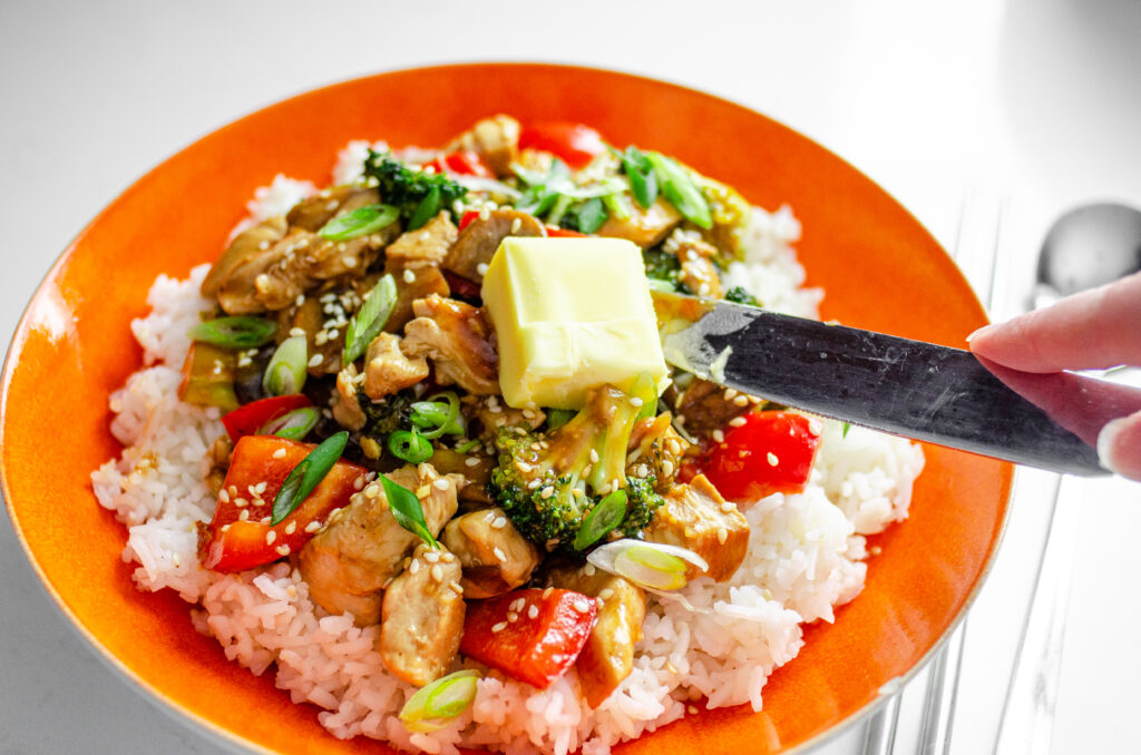 Adding a tablespoon of grass-fed butter to a bowl of healthy chicken stir fry on top of white rice.