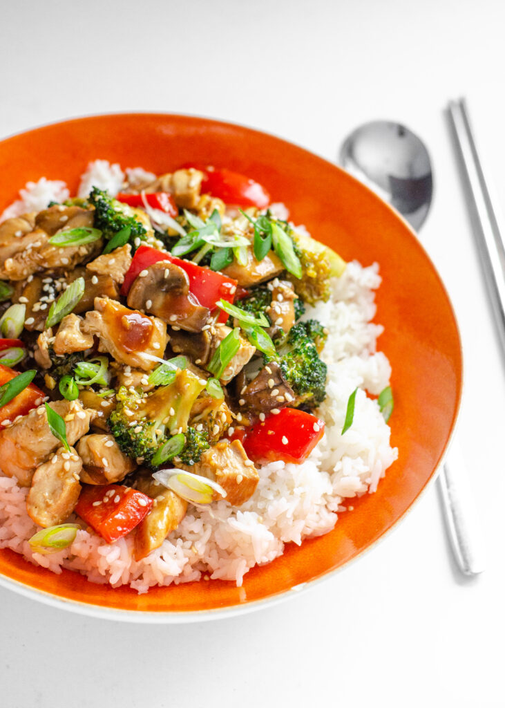 A bowl of healthy chicken stir fry on top of a bed of rice with a spoon and chopsticks next to it.