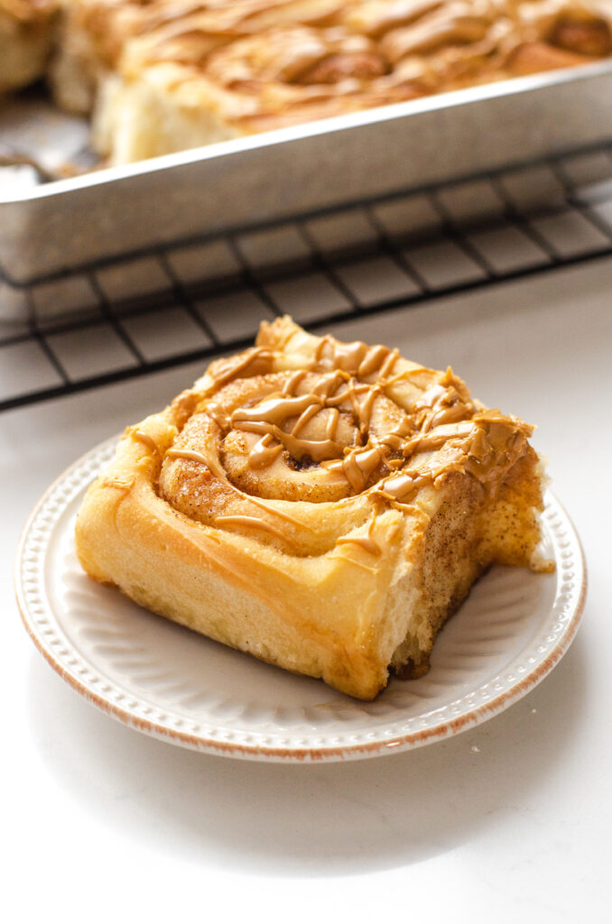 A dairy free cinnamon roll on a plate with the pan of cinnamon rolls in the background. 