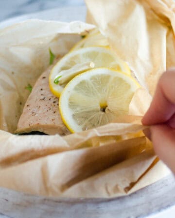 A salmon filet in a piece of parchment paper with snow peas underneath and slices of lemon on top.