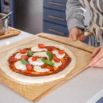 Transferring the topped sourdough pizza crust onto a pizza peel to transfer to the oven.