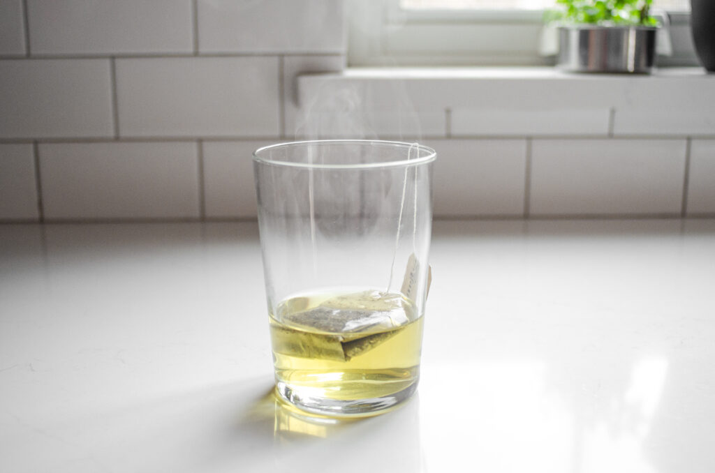 A tea bag and hot water in a drinking glass on a white countertop.