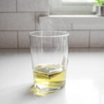 A tea bag and hot water in a drinking glass on a white countertop.