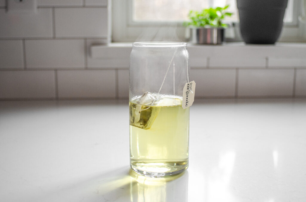 AA tea bag and hot water in a drinking glass on a white countertop.