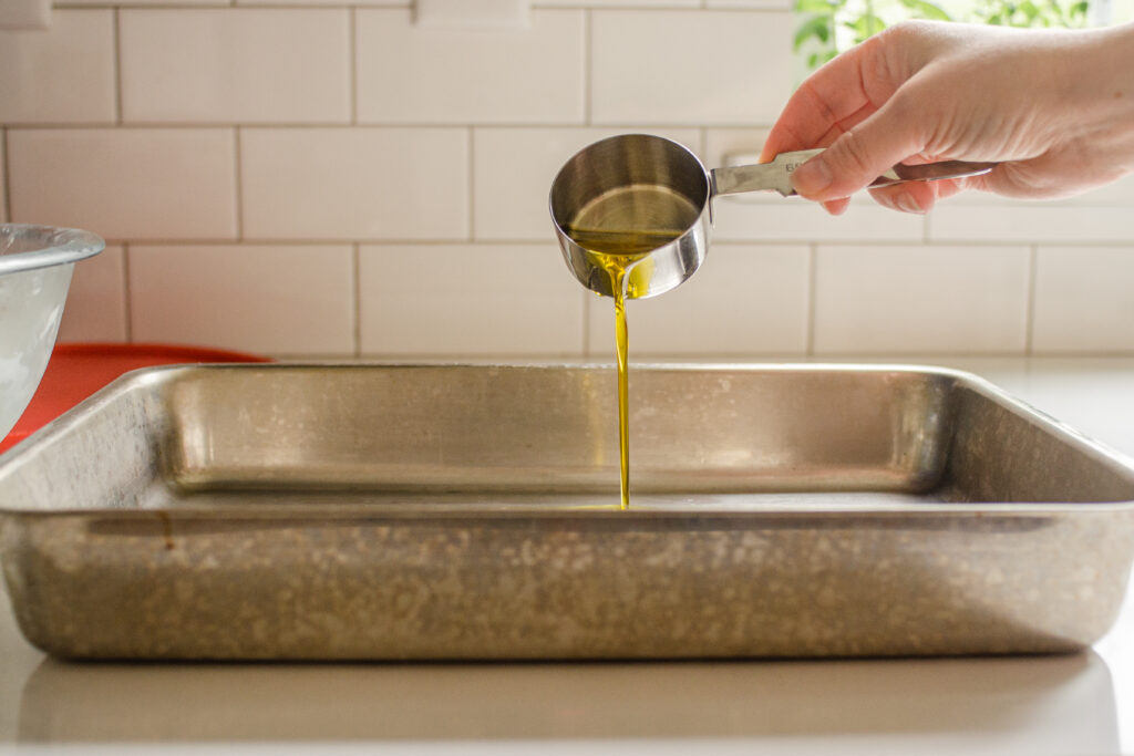 Pouring extra virgin olive oil into a pan to grease it in preparation for putting the focaccia dough in it.