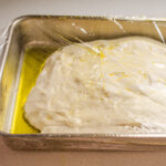 The sourdough focaccia rising in the pan with plastic wrap on top.