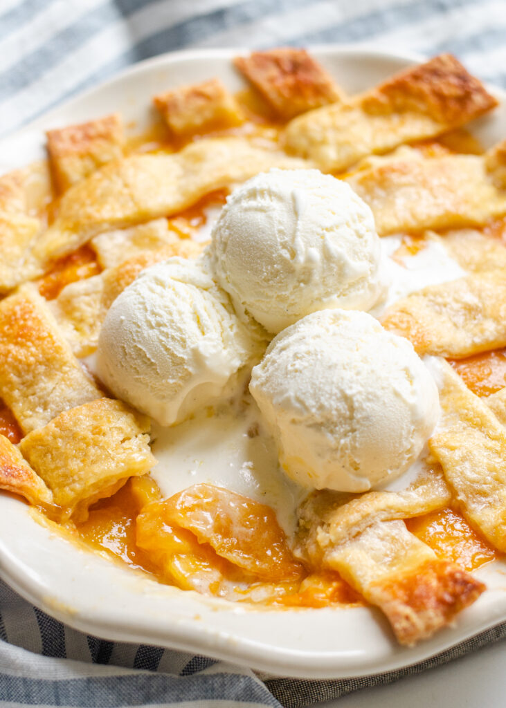 Southern Peach cobbler in a pie dish with a lattice pie crust topping with three scoops of vanilla bean ice cream on top.