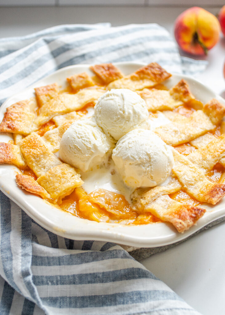 Southern Peach cobbler baked in a pie dish with a lattice pie crust topping and scoops of vanilla bean ice cream on top.