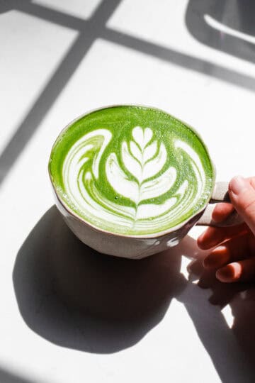 Holding a mug of homemade Starbucks hot matcha green tea latte on a sunny counter.
