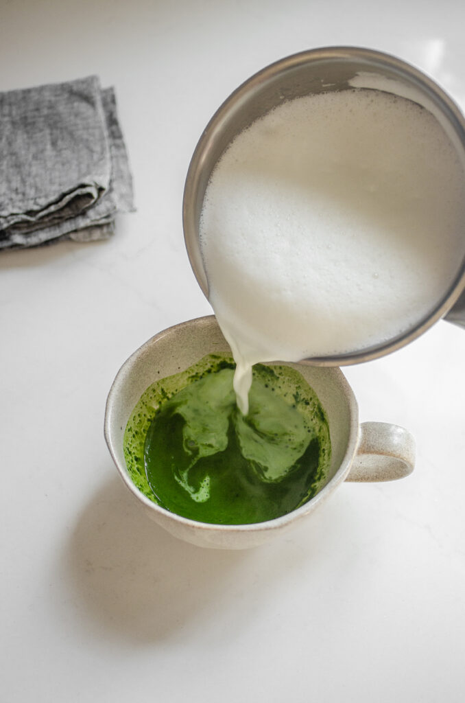 Pouring the frothed milk over the whisked matcha in the mug.