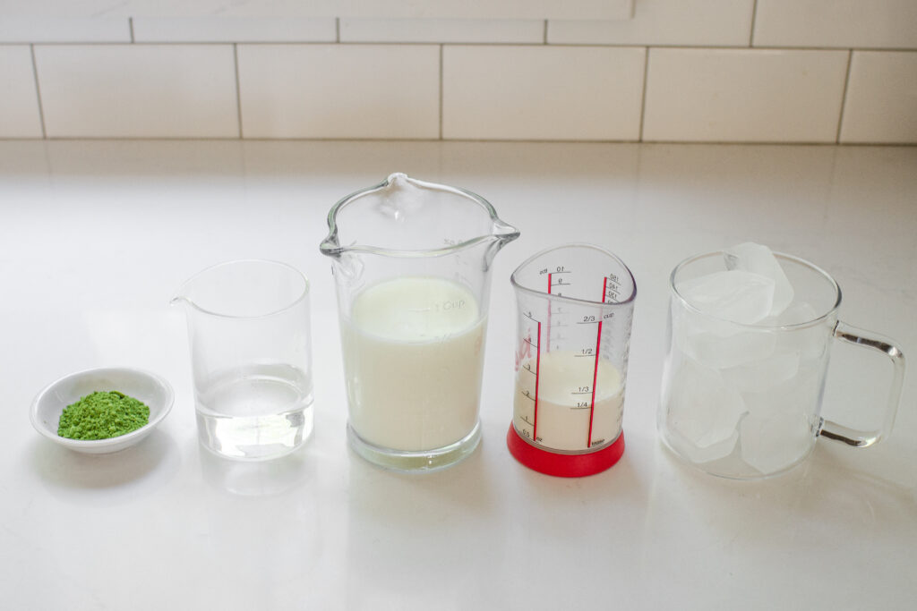 All of the ingredients needed to make matcha milk tea laid out on a white countertop.