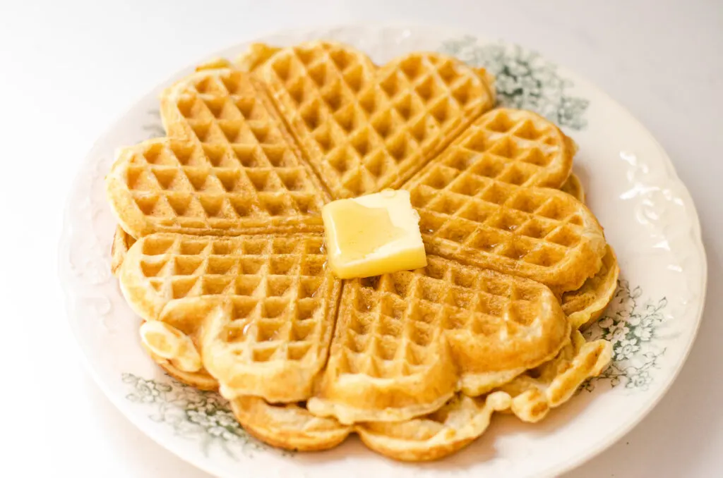 A cooked sourdough waffle on a vintage plate with a pat of butter and maple syrup on top.