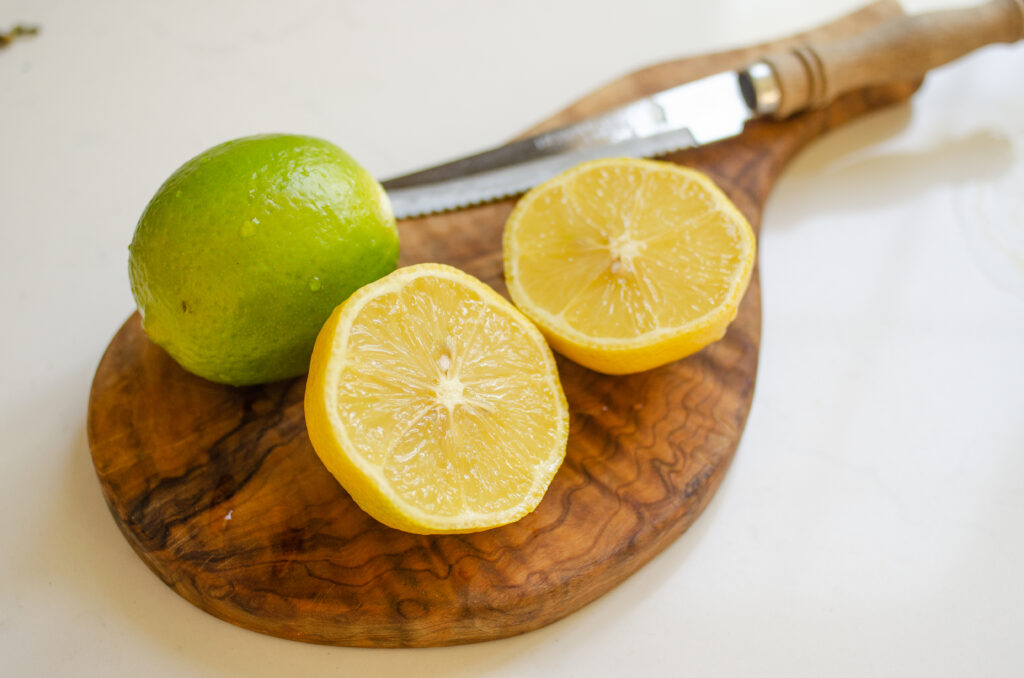 A lime on a cutting board with a lemon next to it cut in half and a serrated knife behind it.