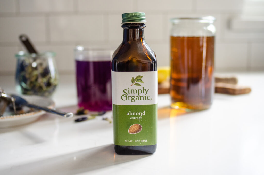 A photo of almond extract with butterfly pea tea in the background along with maple syrup. 