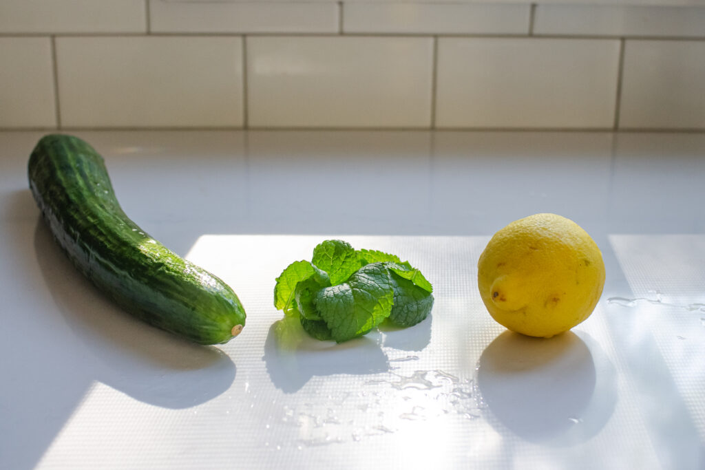 A cucumber, mint leaves, and lemon on a white countertop with the sunlight shining through the window.