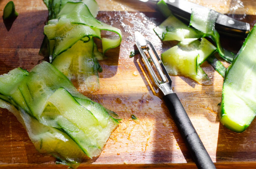 Long cucumber peels on a wooden cutting board with a vegetable peeler off to the side.