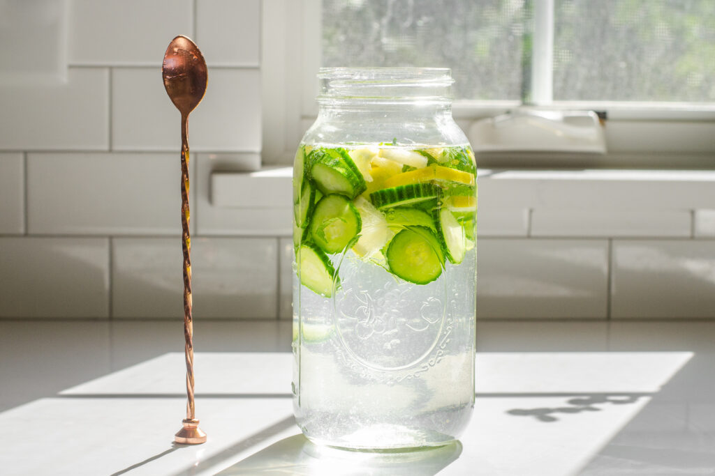 A large mason jar with lemon, cucumbers, and mint on a sunny countertop with a stirring spoon off to the side.