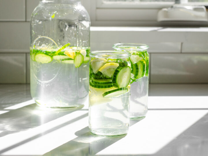 Two glasses of cucumber lemon mint water on a white countertop with the sunlight coming through the window behind them.
