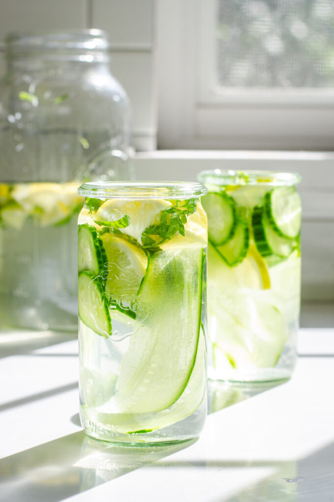 Two glasses of cucumber lemon mint water on a white countertop with the sunlight coming through the window behind them.