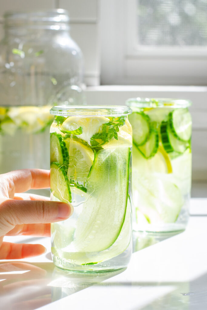 Two glasses of cucumber lemon mint water on a white countertop with the sunlight coming through the window behind them.