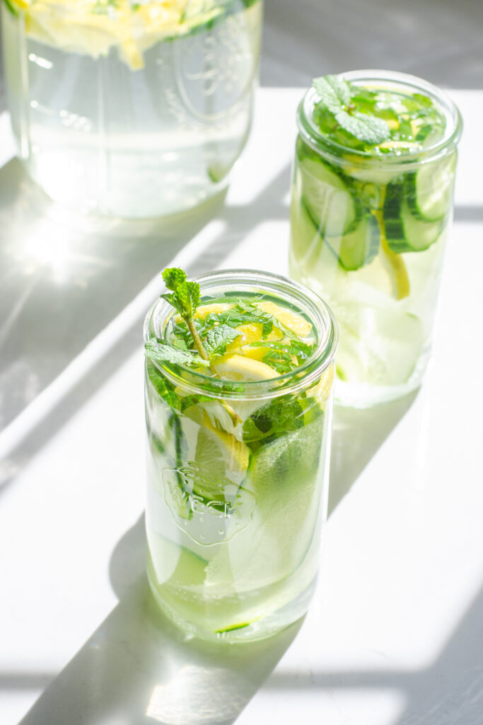 Two glasses of cucumber lemon mint water on a white countertop with the sunlight coming through the window behind them.