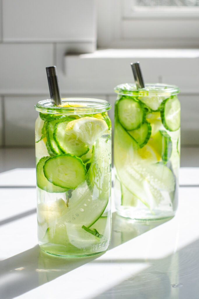 Two glasses of cucumber lemon mint water on a white countertop with the sunlight coming through the window behind them.