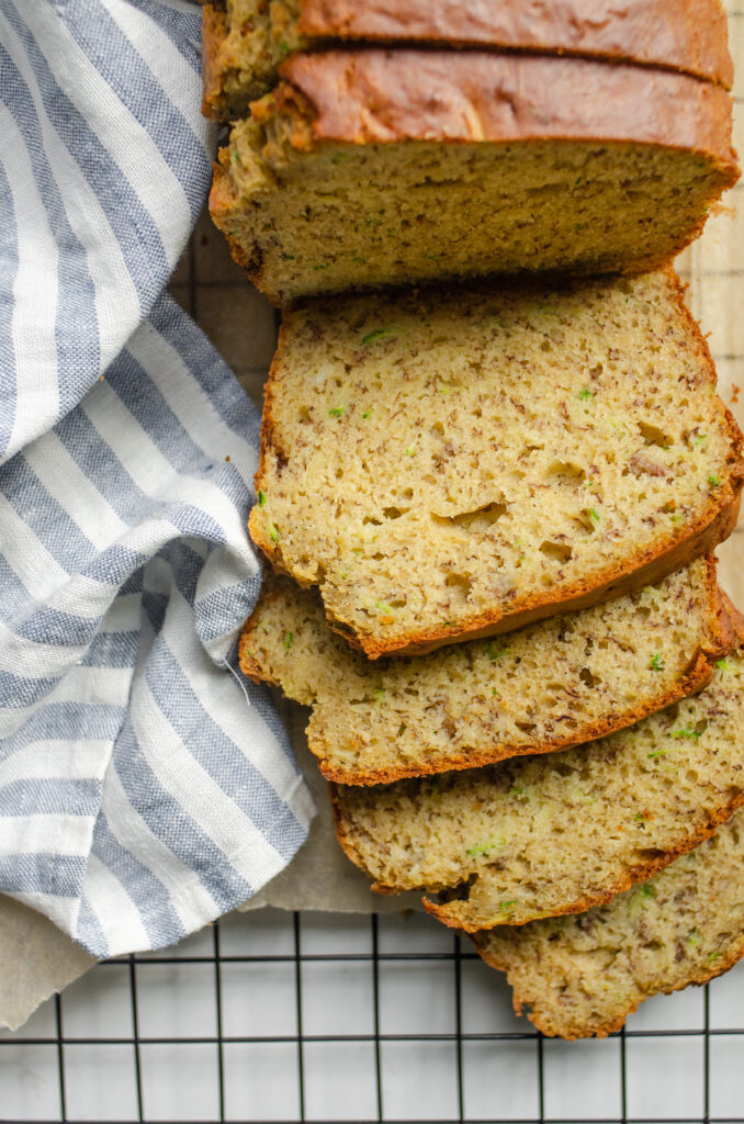 Slices of banana zucchini bread on top of parchment paper on top of a wire cooling rack with a linen striped tea towel off to the side.