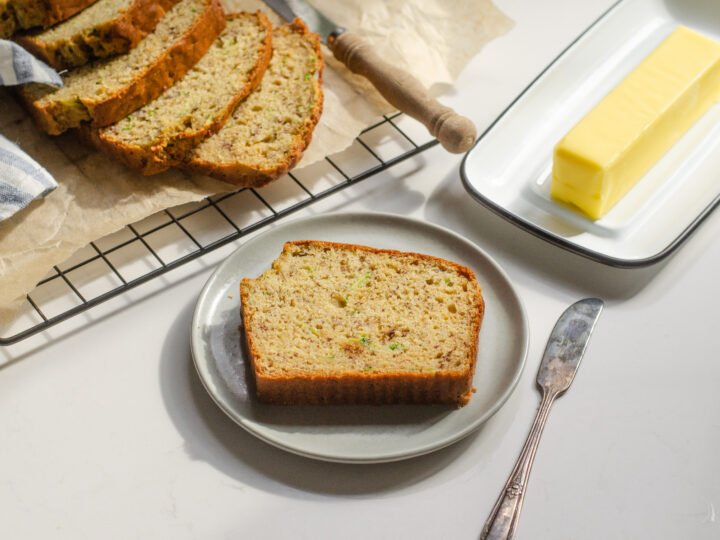 A slice of banana zucchini bread on a plate with the rest of the loaf behind it and butter off to the side.