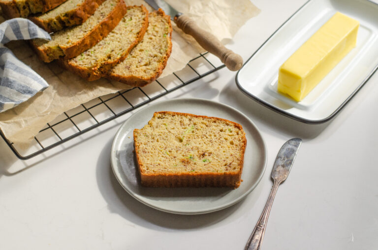 A slice of banana zucchini bread on a plate with the rest of the loaf behind it and butter off to the side.
