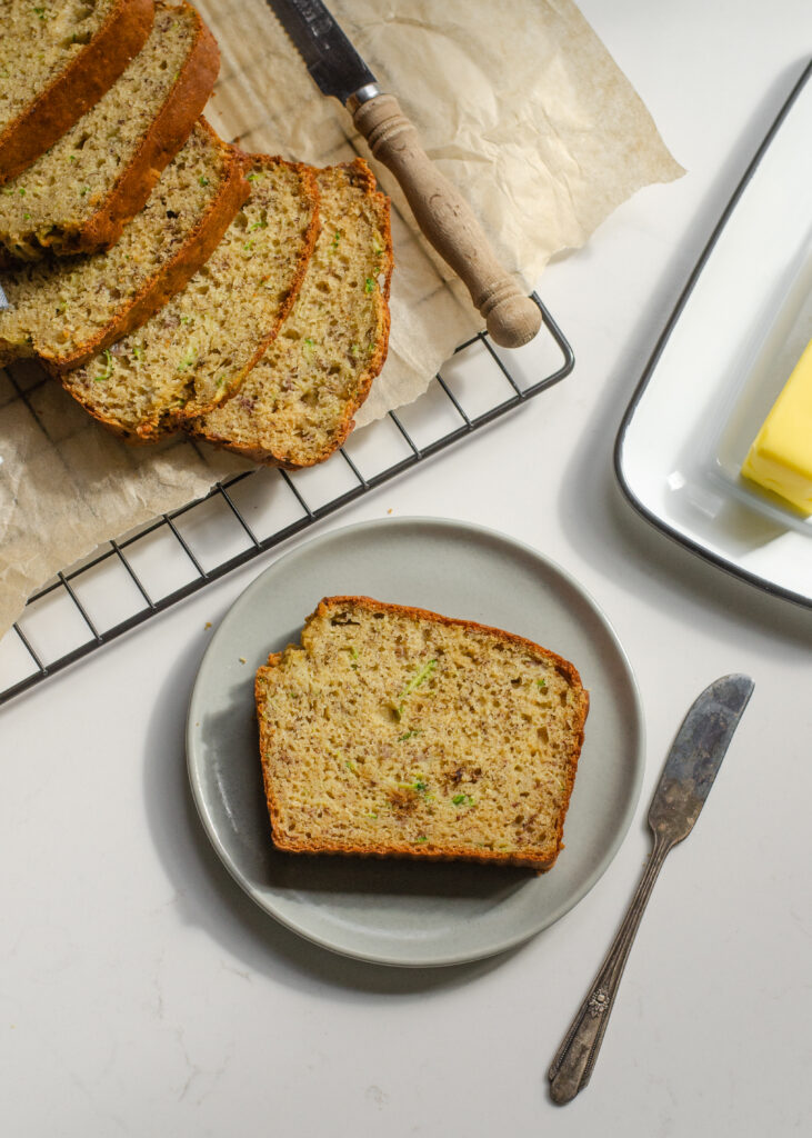 A slice of banana zucchini bread on a plate with the rest of the loaf behind it and butter off to the side.