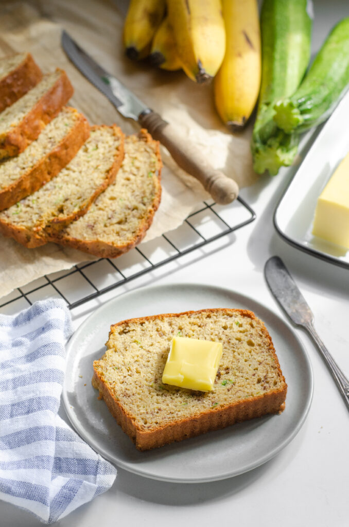 A slice of banana zucchini bread on a plate with the rest of the loaf behind it and butter off to the side.