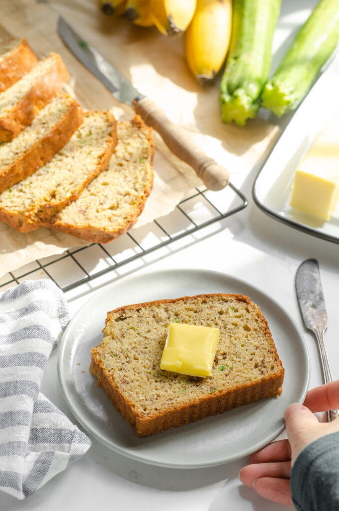 A slice of banana zucchini bread on a plate with the rest of the loaf behind it and butter off to the side.