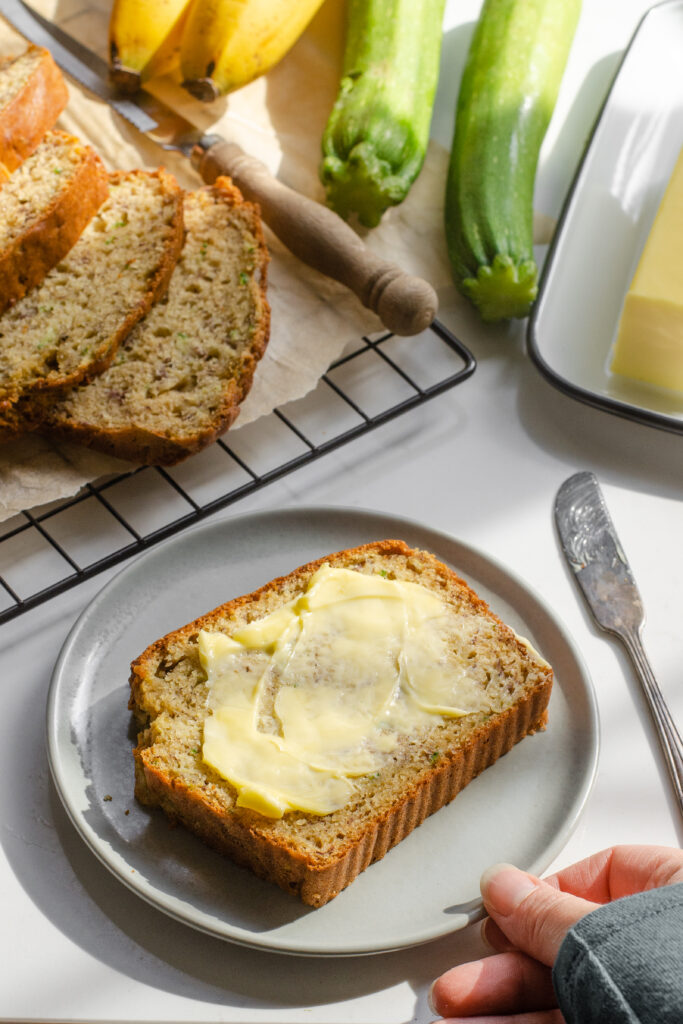 A slice of banana zucchini bread on a plate with the rest of the loaf behind it and butter off to the side.