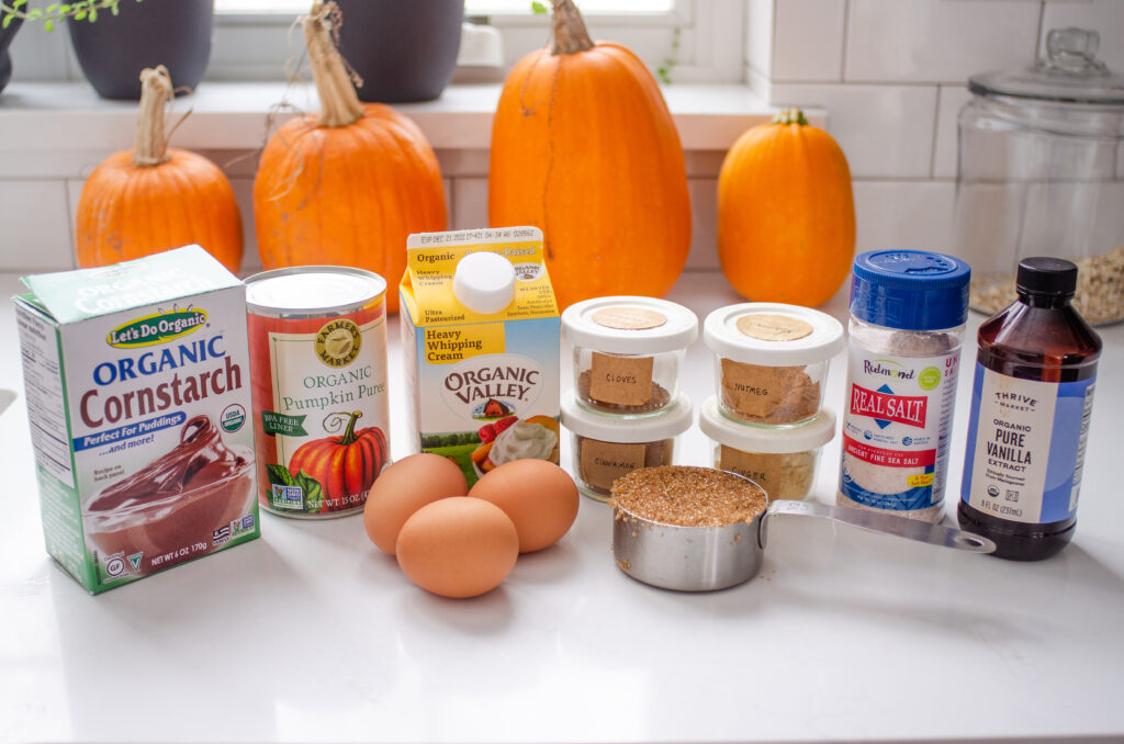 All of the ingredients needed to make a low sugar pumpkin pie at home laid out on a white countertop with fresh pumpkins in the background. 