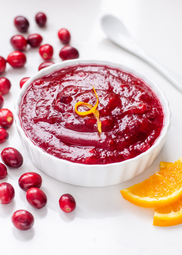 A ramekin of organic cranberry sauce with fresh cranberries, orange slices, and a white spoon around it.