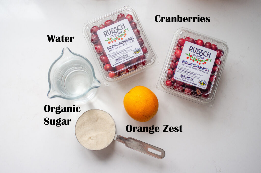 All of the ingredients necessary to make organic cranberry sauce laid out on a white countertop with words labeling each ingredient.