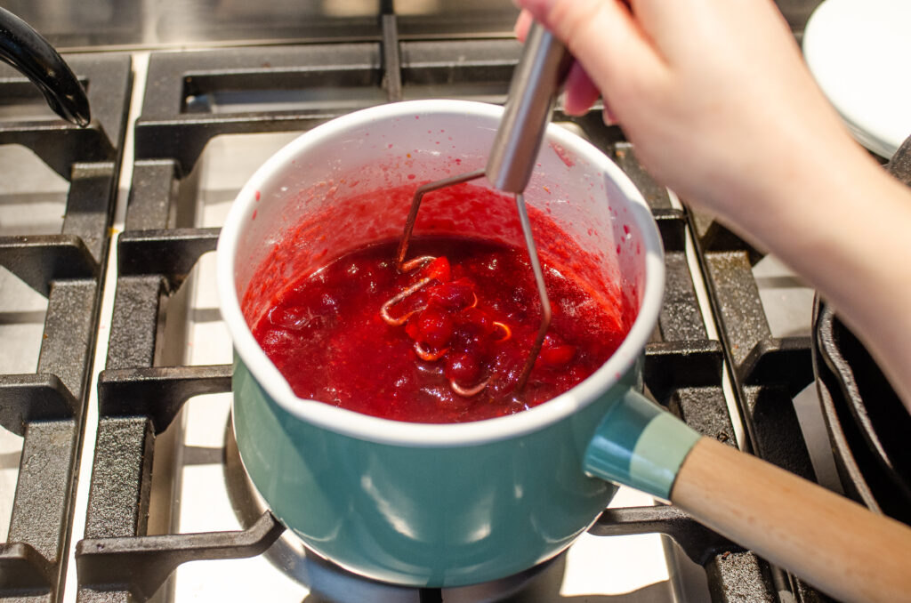 Mashing the cranberry sauce in the saucepan with a mini potato masher to break up the cranberries.
