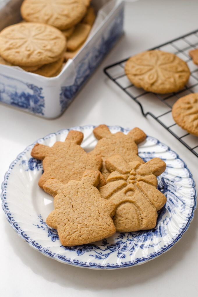 Speculoos cookies on a blue and white plate with more cookies in a tin behind them, and on a wire rack to the side.