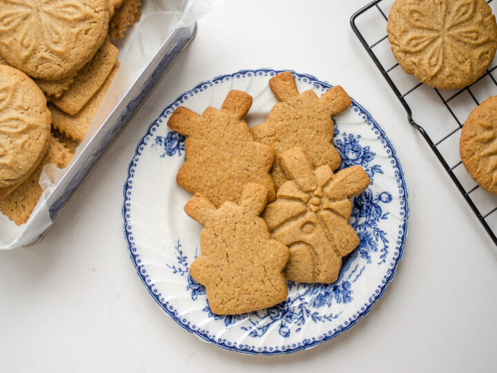 Speculoos cookies on a blue and white plate with more cookies in a tin behind them, and on a wire rack to the side.