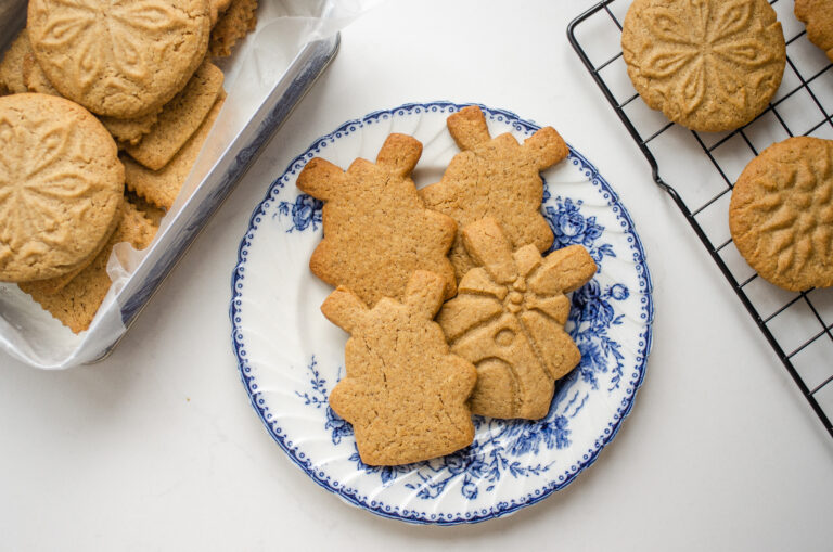 Speculoos cookies on a blue and white plate with more cookies in a tin behind them, and on a wire rack to the side.