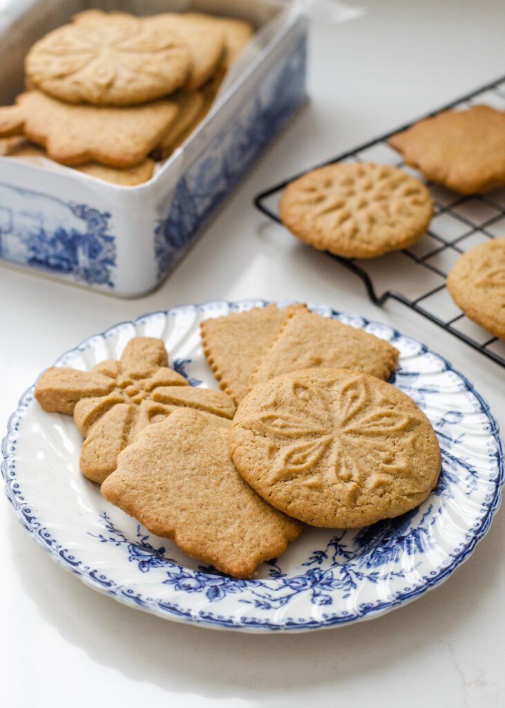 Speculoos cookies on a blue and white plate with more cookies in a tin behind them, and on a wire rack to the side.